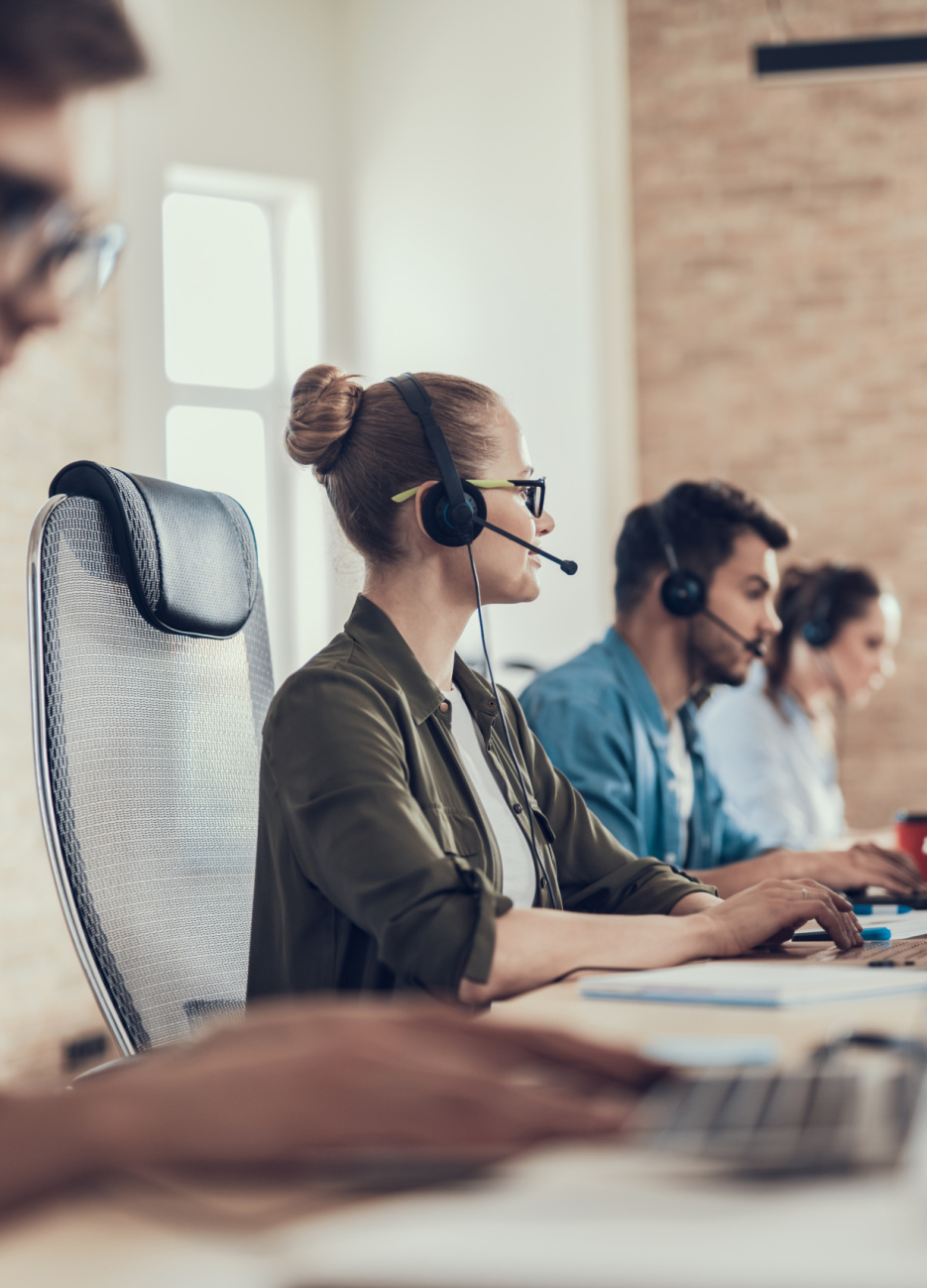 Waist up portrait of smiling woman sitting in call center while looking computer screen while using headset with colleagues on background